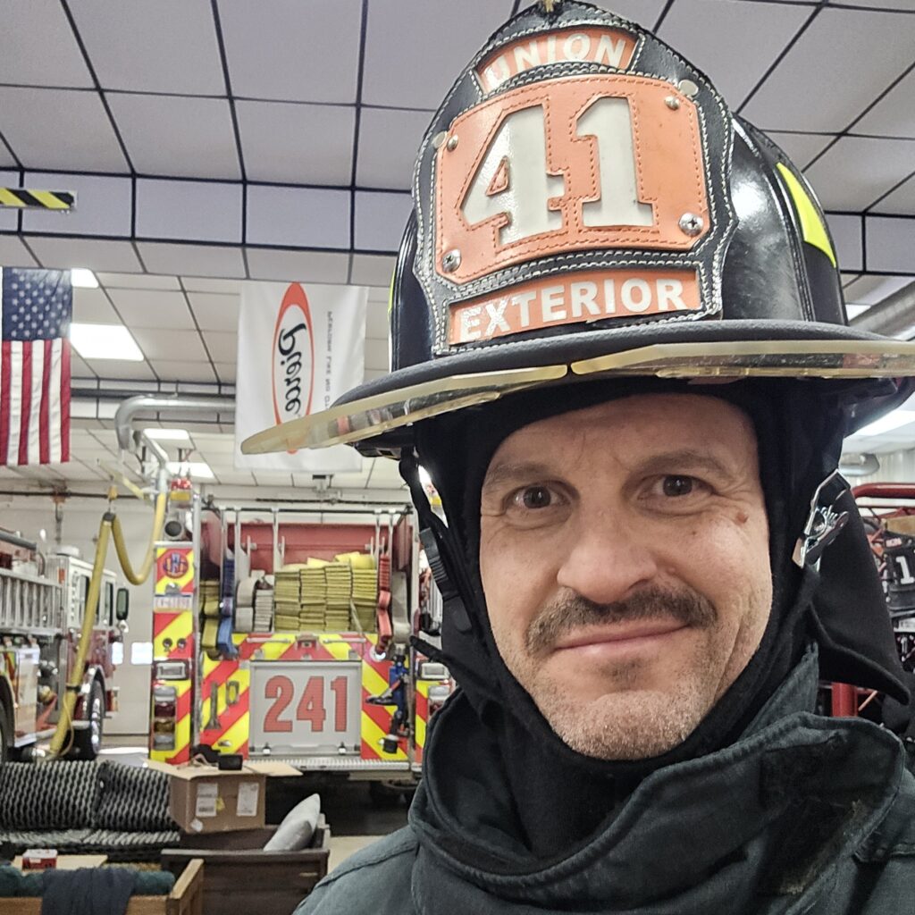 about me image of derek bicksler, volunteer firefighter at Carlisle Union 41, in turnout gear and helpmet standing in garage bay at union 41 station