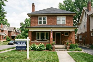 A photo of a historic all brick with front porch 1920 era single family home with a generic dark blue for sale sign in the yard. similar homes surround it (but they are different but of same era)